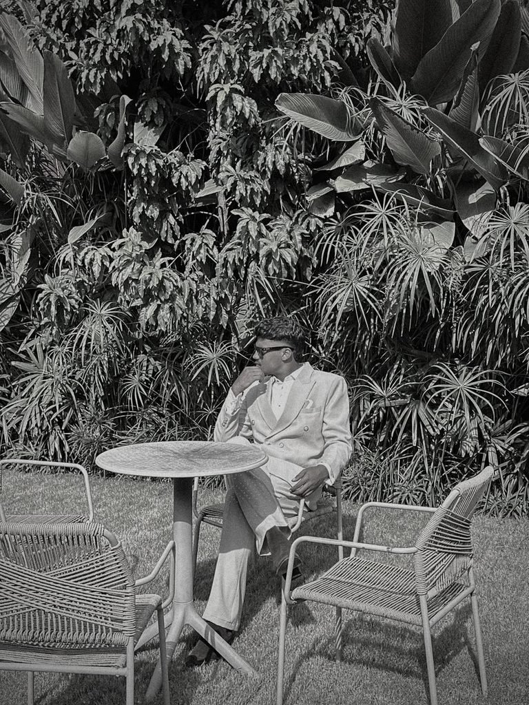 A professional portrait of a freelance digital marketer in Malappuram wearing a stylish white suit and sunglasses, seated in a beautiful outdoor garden setting. The image represents a modern, professional, and confident personal brand.
